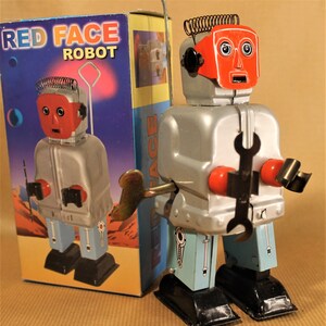May include: A silver wind-up robot toy with a red face and a spanner in its hand. The robot is standing on a black base and has a blue and silver body. The box reads "Red Face Robot".