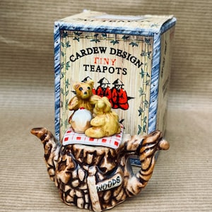 May include: A brown ceramic teapot shaped like a tree stump with a red and white checked picnic blanket on top. Three teddy bears are sitting on the blanket, one holding a cupcake. The teapot has a label that says "Woods". The box the teapot is in says "Cardew Design Tiny Teapots".