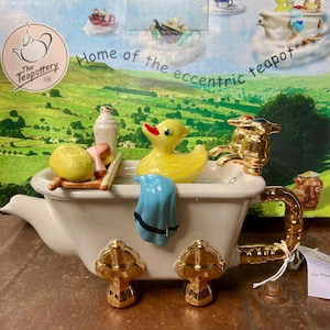May include: A ceramic teapot shaped like a white clawfoot bathtub with gold-coloured feet and handle. It features a yellow rubber duck, a blue towel, a white bottle, and a sponge inside. The faucet is gold-coloured. The box reads The Teapottery.