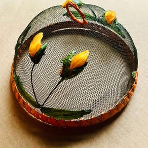 May include: A decorative food cover with a black mesh dome and a woven orange and brown rim. The cover is adorned with yellow flower buds and green leaves, and has a brown handle. The overall design is floral.