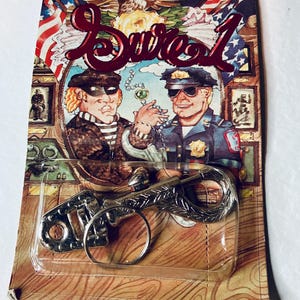 May include: A vintage key ring in a clear plastic package. The key ring is silver-coloured and shaped like a gun. The packaging features a cartoon illustration of a police officer and a burglar, with the word "Sure 1" in a burgundy script.