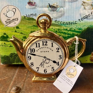 May include: A gold-coloured ceramic teapot shaped like a round clock with a white face, black numerals, and the text "Everything stops for tea". It features a top handle, a side spout, and a white paper tag attached to the handle with a string.