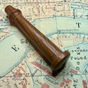 May include: A cylindrical wooden object with a smooth, polished brown finish, featuring a flared base and a rounded, tapered top section. The item has a consistent grain pattern throughout its length and is positioned diagonally across the frame.