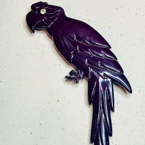 May include: A dark purple parrot-shaped brooch with a crystal accent on its head. The brooch is carved with detailed feather and wing patterns. The parrot's beak is slightly open, and its tail feathers are long and pointed.