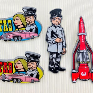 May include: A set of wooden cutouts depicting characters and vehicles from the classic TV show, Thunderbirds. Includes a pink car with the word "FAB", a red rocket, and a character in a gray suit.