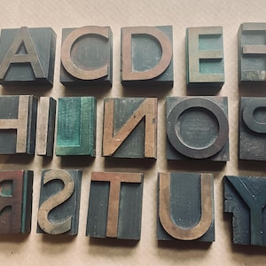 May include: A selection of vintage wooden letterpress printing blocks. The blocks display uppercase letters A, C, D, E, F, H, I, N, O, P, R, S, T, U, and Y. The letters are in a mix of dark green and brown tones.