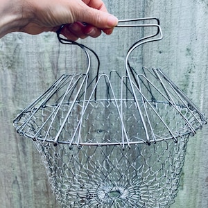 May include: A collapsible silver-toned metal wire basket featuring a flexible mesh structure that expands and contracts. The basket includes a central handle mechanism made of rigid wire loops that allow for easy lifting and folding of the unit.
