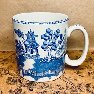May include: White ceramic mug with a blue Willow pattern design. The mug features a handle and a detailed landscape scene with a pagoda, trees, and a fence. The rim of the mug has a blue dotted pattern.