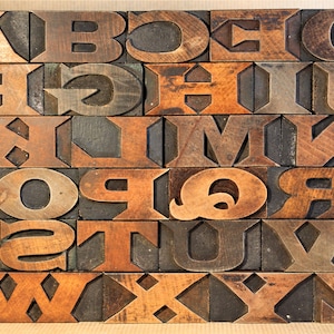 May include: A set of wooden letterpress blocks, each with a different letter of the alphabet, arranged in alphabetical order. The blocks are made of dark brown wood and have a worn, vintage look.