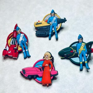 May include: Set of five colourful plastic figurines of characters from the TV show Thunderbirds. Each figurine is standing on a base with a vehicle from the show. The vehicles include a red rocket, a yellow truck, a pink car, a green spaceship, and a blue jet.