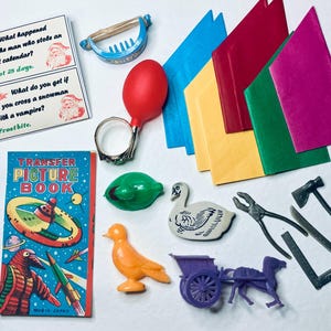 May include: A selection of vintage toys and ephemera, including a Transfer Picture Book, joke cards, and various plastic figurines. The colour palette includes red, blue, green, and yellow. There is also a red bulb, a ring, and a swan figurine.
