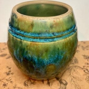 May include: A handcrafted ceramic vase with a rounded barrel shape. The vase features a blend of green and teal glazes, with a band of teal around the middle. The interior is a deep green. The vase is a decorative item.