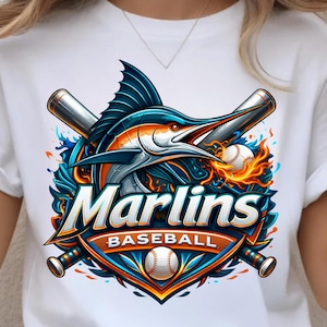 May include: White t-shirt featuring a colorful graphic of a marlin fish with crossed baseball bats, a baseball, and the text "Marlins Baseball". The design incorporates shades of blue, orange, and silver.