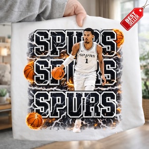 Pu&ograve; includere: T-shirt bianca con un giocatore di basket in uniforme Spurs, e la parola "SPURS" in un audace design in bianco e nero. Il design include palloni da basket ed effetti fulmine.