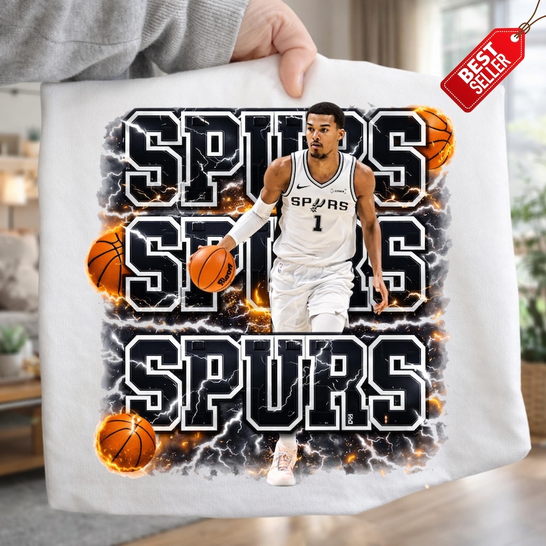 Pu&ograve; includere: T-shirt bianca con un giocatore di basket in uniforme Spurs, e la parola "SPURS" in un audace design in bianco e nero. Il design include palloni da basket ed effetti fulmine.