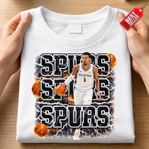 Pu&ograve; includere: Felpa bianca girocollo con grafica di un giocatore di basket in uniforme Spurs, con la scritta "SPURS" in grassetto e bordata. Sono inclusi anche palloni da basket ed effetti di fulmini.