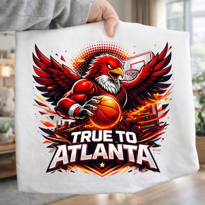 May include: White t-shirt featuring a red eagle graphic with a basketball, wings spread, and the text "TRUE TO ATLANTA." The design includes a basketball hoop and fiery accents, creating a dynamic sports-themed image.
