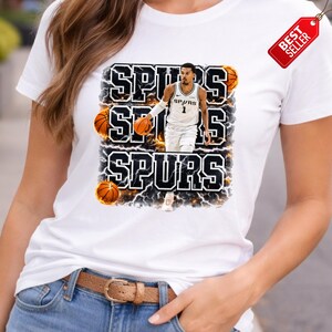 Pu&ograve; includere: T-shirt bianca con grafica di un giocatore di basket in uniforme Spurs, con la scritta "SPURS" in grassetto e contornata. Il design include palloni da basket ed effetti fulminei. &Egrave; presente un'etichetta "Best Seller".