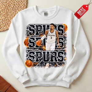 Pu&ograve; includere: Felpa bianca con un giocatore di basket in uniforme Spurs, e la parola "SPURS" impilata tre volte in un carattere in grassetto e sottolineato. Grafiche di palloni da basket e fulmini sono incorporate nel design. Un'etichetta "Pi&ugrave; venduto" &egrave; visibile.