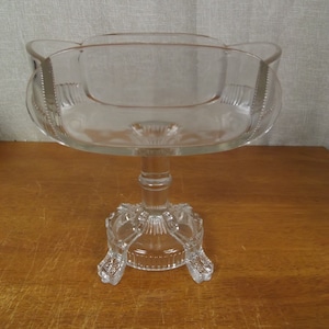 May include: A clear glass pedestal bowl with a scalloped rim and a decorative base. The bowl is on a wooden surface.