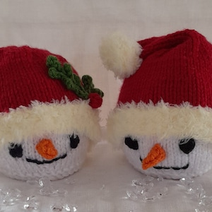 以下が含まれることがあります： 赤いサンタの帽子をかぶったニットの雪だるまのオーナメントが2つ。白いファーのトリムと黒い目があります。一方の雪だるまには緑のヒイラギの枝があり、もう一方の雪だるまには帽子の先に白いポンポンが付いています。