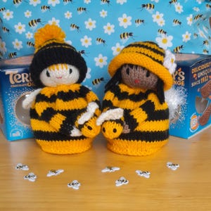 May include: Two knitted bee dolls, each in yellow and black striped outfits and matching hats. One doll has a black hat with a yellow bobble, the other a yellow hat with a white flower. Small bee-shaped decorations are scattered in front of the dolls.
