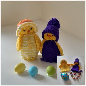 May include: Two crocheted chick toys, one wearing a yellow and white striped outfit with an orange hat and the other wearing a purple outfit with a purple hat. Three pastel coloured eggs and a chocolate egg are in the foreground.
