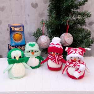 Puede incluir: Cuatro adornos de pingüinos tejidos a mano en verde, rojo y blanco, con gorros y bufandas a juego. Un pequeño árbol de Navidad y dos adornos decorativos en el fondo. También hay una caja de Terry's Orange.