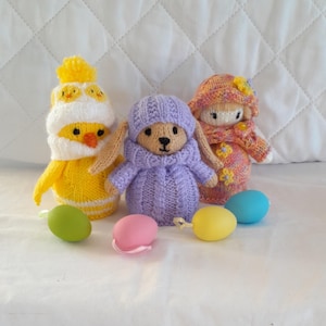 May include: Three knitted Easter figures: a yellow chick, a lavender bunny, and a doll in a pink and orange outfit. The chick wears a white hat with yellow accents. The bunny has long ears. Colourful Easter eggs are in front.