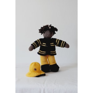 Może przedstawiać: Ręcznie robiona dzianinowa lalka przebrana za strażaka. Lalka ma na sobie granatową kurtkę z żółtymi paskami, żółte spodnie i czarne buty. Obok lalki znajduje się pasujący żółty kapelusz z kwiatkiem. Lalka ma brązowe włosy i neutralny odcień skóry.