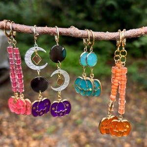 Op de afbeelding: Vier paar Halloween-oorbellen met verschillende ontwerpen, waaronder pompoenen, manen en kralen. De oorbellen zijn gemaakt van goud en zilver en hebben verschillende kleuren, waaronder roze, paars, turquoise en oranje.