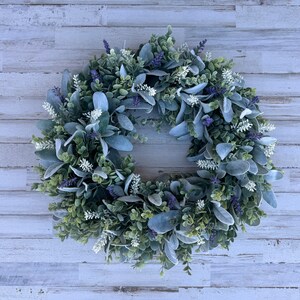 May include: A circular wreath composed of green boxwood foliage, soft grey-green lamb's ear leaves, small white floral sprigs, and clusters of purple lavender-like flowers. The arrangement creates a dense, textured botanical display.