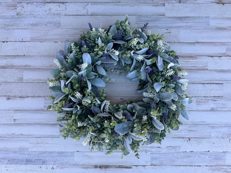 May include: A circular wreath composed of green boxwood foliage, soft grey-green lamb's ear leaves, small white floral sprigs, and clusters of purple lavender-like flowers. The arrangement creates a dense, textured botanical display.
