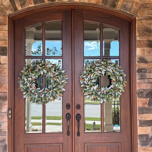 May include: Two identical circular wreaths featuring a mix of sage green lamb's ear leaves and small orange berries are mounted on a set of dark brown wooden double doors with glass panels, framed by a rustic stone archway entrance.