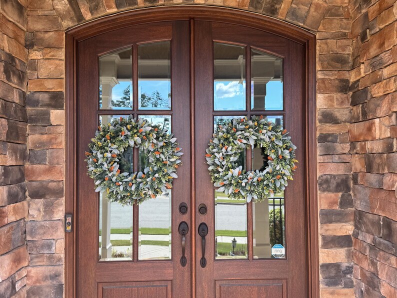 May include: Two identical circular wreaths featuring a mix of sage green lamb's ear leaves and small orange berries are mounted on a set of dark brown wooden double doors with glass panels, framed by a rustic stone archway entrance.