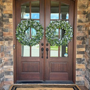 May include: Two round wreaths made of green and silver-toned lamb's ear leaves are hung on a set of dark brown wooden double doors. Below the doors, a rectangular tan coir doormat features a black border and a large letter W in the center.