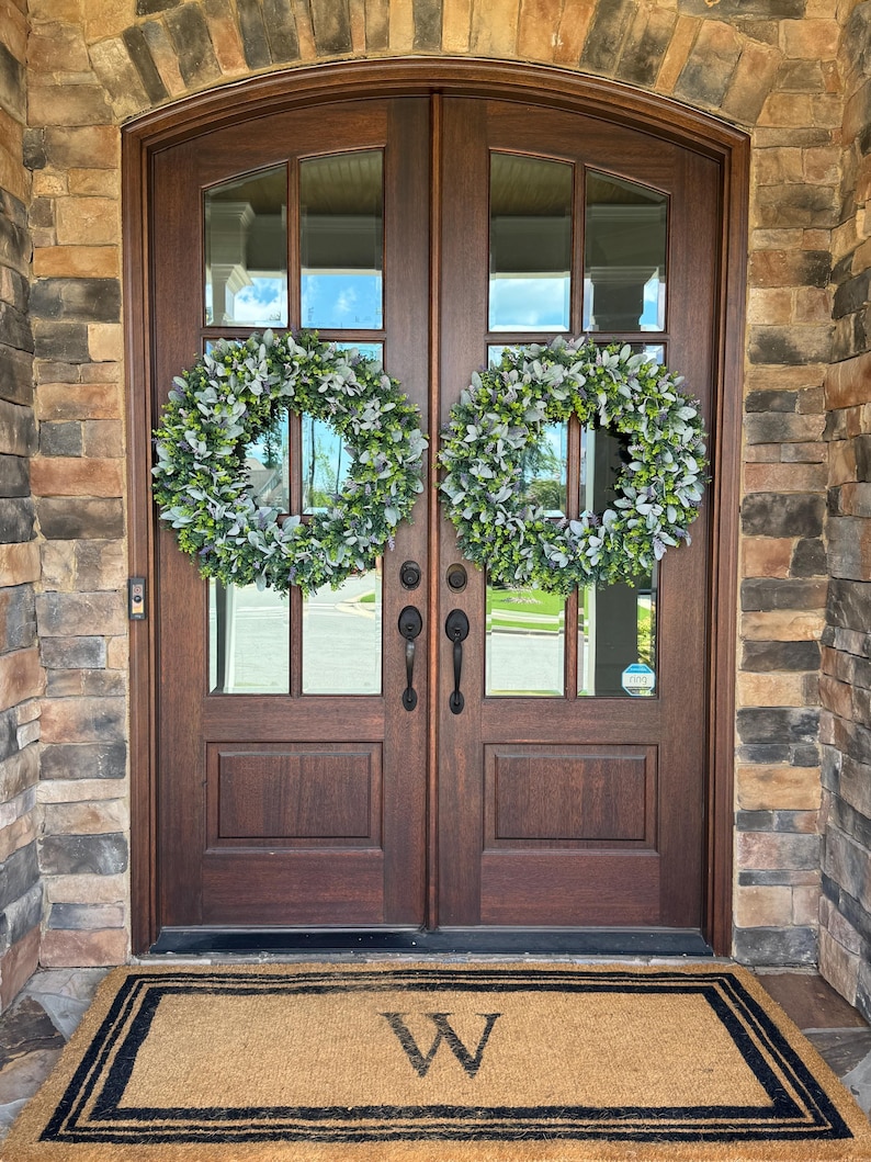 May include: Two round wreaths made of green and silver-toned lamb's ear leaves are hung on a set of dark brown wooden double doors. Below the doors, a rectangular tan coir doormat features a black border and a large letter W in the center.