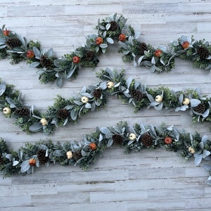 May include: Four strands of artificial greenery garland with faux pine cones, small white pumpkins, and small orange pumpkins. The garland is displayed on a white wood background.