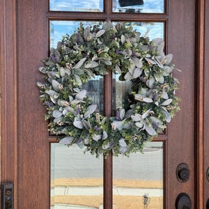 May include: A circular wreath composed of sage green lamb's ear leaves, small dark green foliage, and sprigs of purple lavender flowers. The wreath is mounted on a dark brown wooden door with glass panels and a black metal door handle and doorbell.
