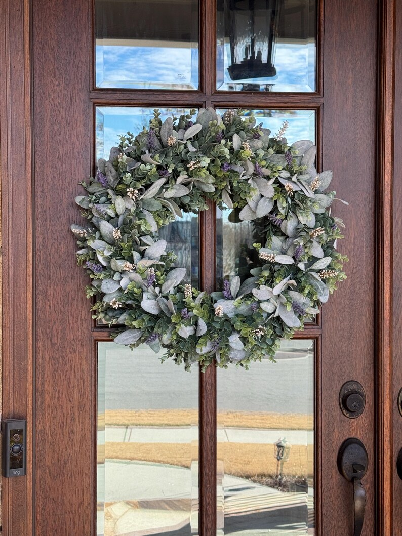 May include: A circular wreath composed of sage green lamb's ear leaves, small dark green foliage, and sprigs of purple lavender flowers. The wreath is mounted on a dark brown wooden door with glass panels and a black metal door handle and doorbell.