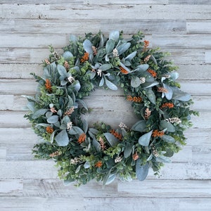 May include: A circular wreath composed of sage green lamb's ear leaves, dark green boxwood foliage, and small clusters of orange and white floral accents. The arrangement features a mix of textures and colors distributed evenly around the ring.