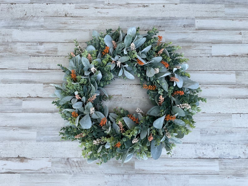 May include: A circular wreath composed of sage green lamb's ear leaves, dark green boxwood foliage, and small clusters of orange and white floral accents. The arrangement features a mix of textures and colors distributed evenly around the ring.
