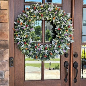 May include: A circular wreath featuring a dense arrangement of green eucalyptus leaves, soft grey lamb's ear foliage, and small clusters of vibrant orange, purple, and white floral accents, mounted on a dark brown wooden double door with glass panels.