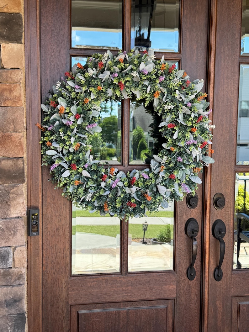 May include: A circular wreath featuring a dense arrangement of green eucalyptus leaves, soft grey lamb's ear foliage, and small clusters of vibrant orange, purple, and white floral accents, mounted on a dark brown wooden double door with glass panels.