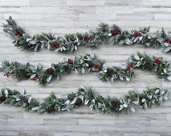 May include: Four strands of eucalyptus garland with red berries and pine cones, displayed on a white wood background.