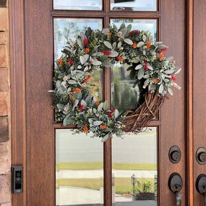 May include: A circular wreath featuring a base of intertwined brown twigs adorned with fuzzy sage green leaves, small clusters of bright orange berries, and tiny white floral accents, mounted on a dark wood door with glass panels.