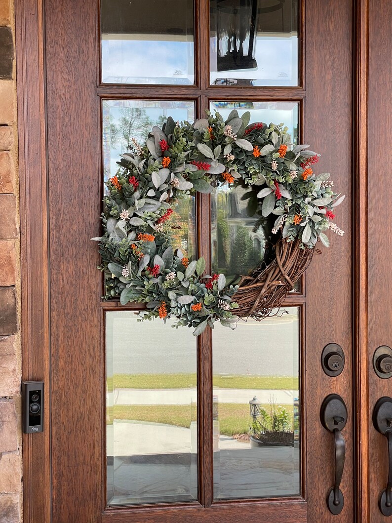 May include: A circular wreath featuring a base of intertwined brown twigs adorned with fuzzy sage green leaves, small clusters of bright orange berries, and tiny white floral accents, mounted on a dark wood door with glass panels.