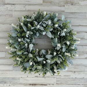 May include: A circular wreath composed of dense green foliage and soft, sage-colored lamb's ear leaves, accented with small, upright white floral sprigs distributed throughout the arrangement to create a textured, layered botanical display.