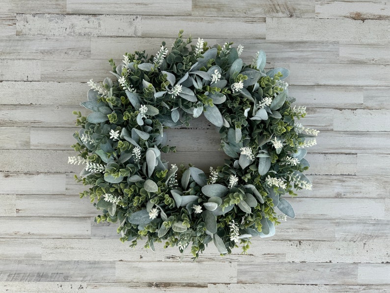 May include: A circular wreath composed of dense green foliage and soft, sage-colored lamb's ear leaves, accented with small, upright white floral sprigs distributed throughout the arrangement to create a textured, layered botanical display.