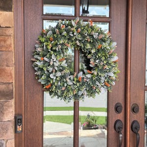 May include: A circular wreath featuring a dense arrangement of green eucalyptus leaves, soft grey lamb's ear foliage, and small clusters of bright orange floral accents, mounted on a dark brown wooden door with glass panels and metal hardware.